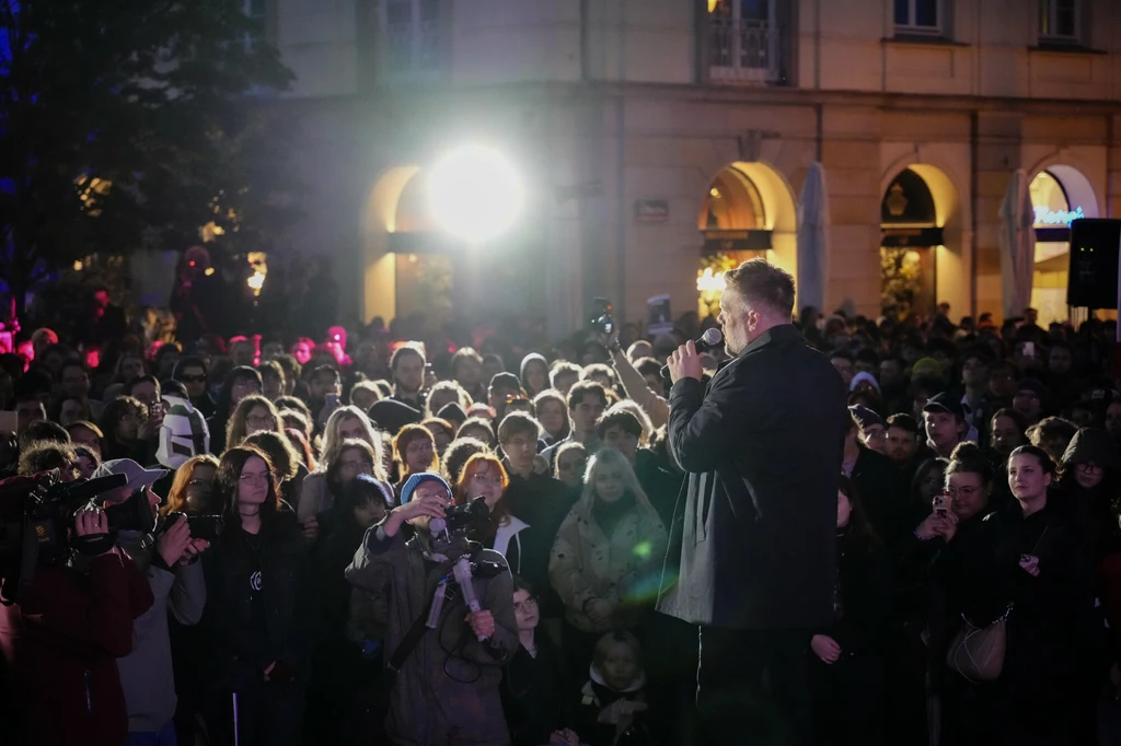 Wiec Adriana Zandberga w Warszawie Mężczyzna przemawiający do tłumu podczas wydarzenia na otwartej przestrzeni, wieczorową porą, w miejskim otoczeniu. W tle oświetlone budynki i jasne reflektory.